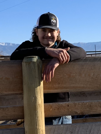  A young man with curly hair wearing a cap leans against a fence post.