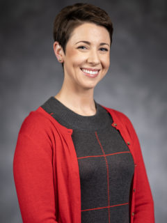 A professional headshot of a smiling woman with short dark hair. She is wearing a cherry colored cardigan over a charcoal and red  wool sweater.