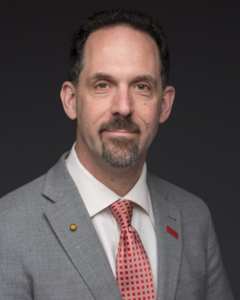 A professional headshot of a man with short dark hair and a van dyke beard. He is wearing a gray suit with a red and tan checked tie.