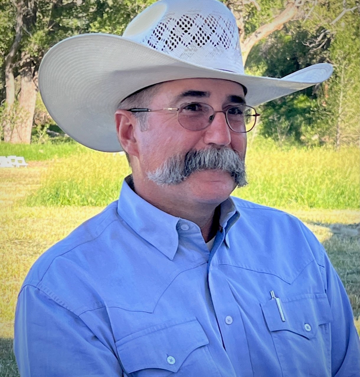 a man with a bushy gray mustache and wire-rimmed glasses is wearing a straw cowboy hat and a blue oxford shirt
